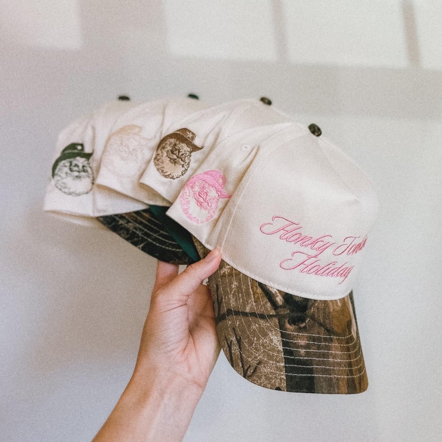 Hand holding a camouflage-patterned cap with floral designs and text against a white background