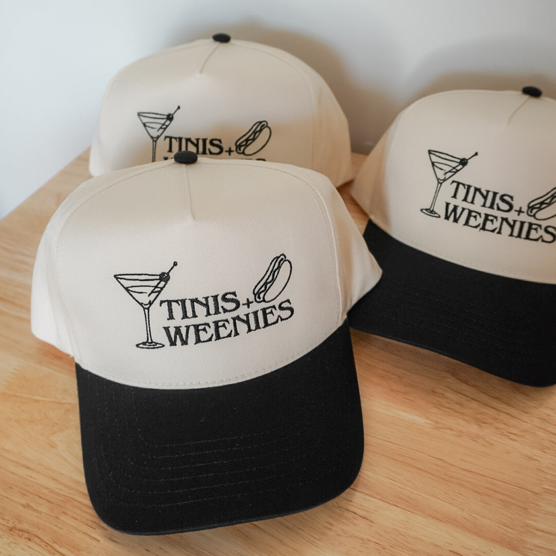 Three baseball caps with 'Tinis & Weenies' logo on a wooden surface.