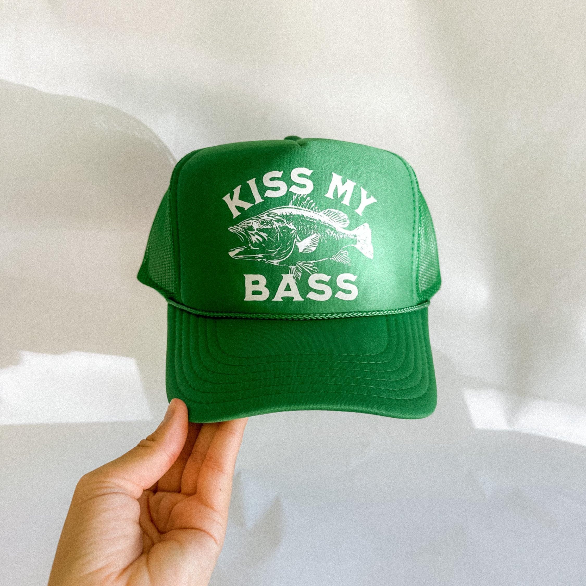 Green cap with 'Kiss My Bass' text and fish graphic held by a hand on a light background