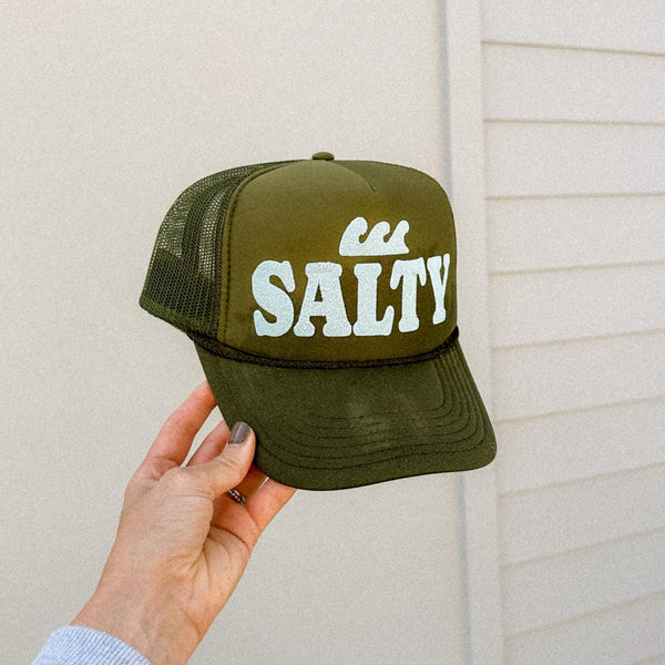 Green cap with 'Salty' text held by a hand against a beige wall.